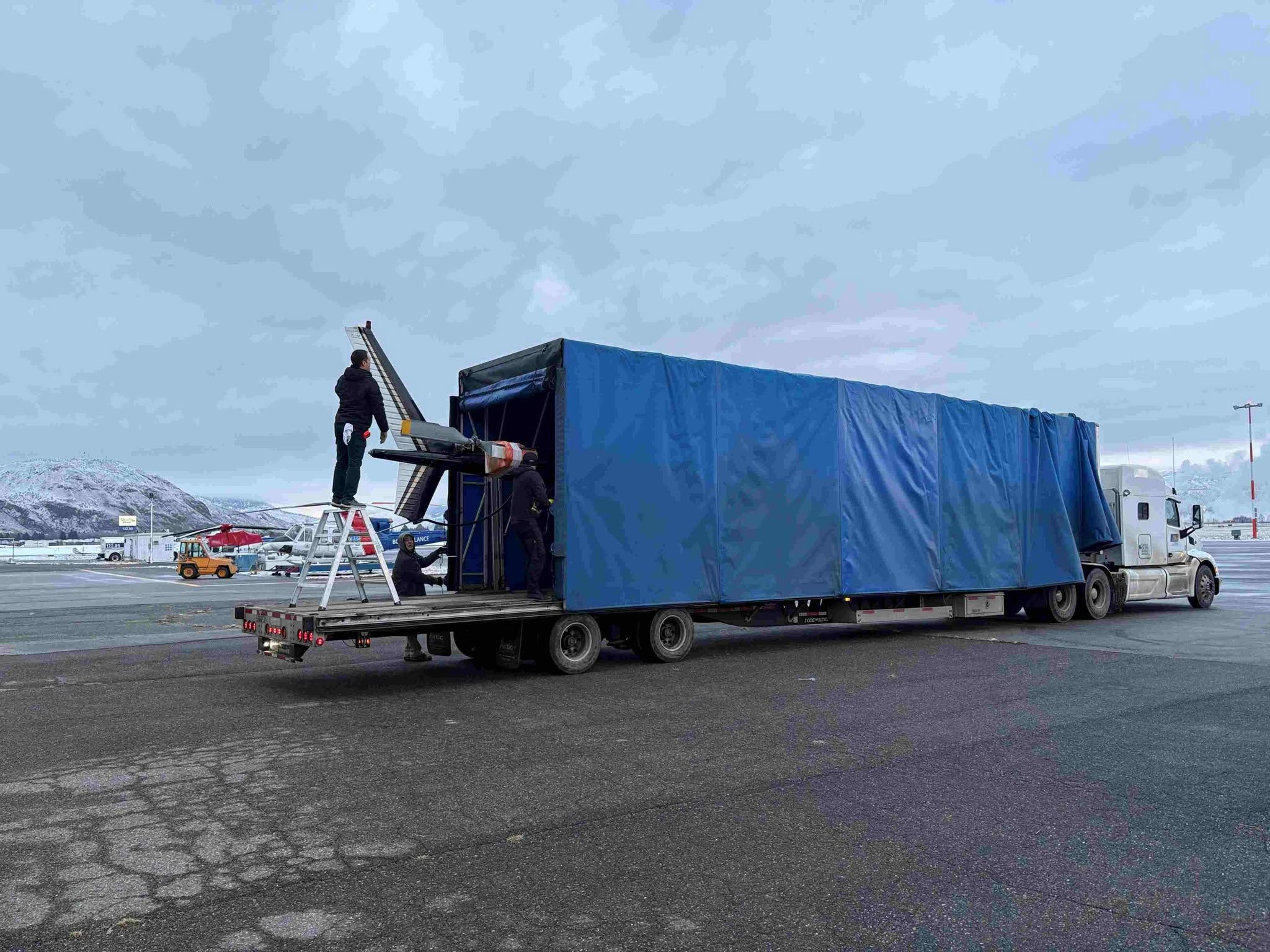 Team loading a helicopter into a covered semi-trailer for secure ground transport at a snowy mountain airport.