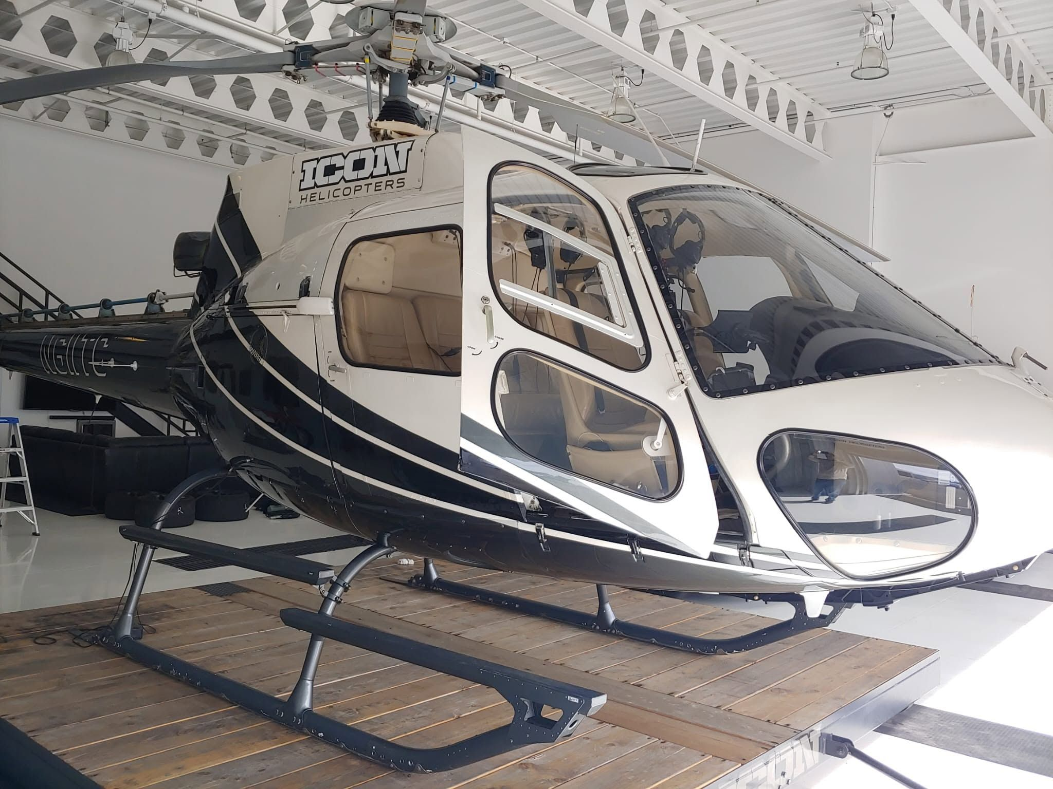 Single-engine helicopter in white and black livery with a beige leather interior, parked on a wooden ground-handling platform inside the hangar.
