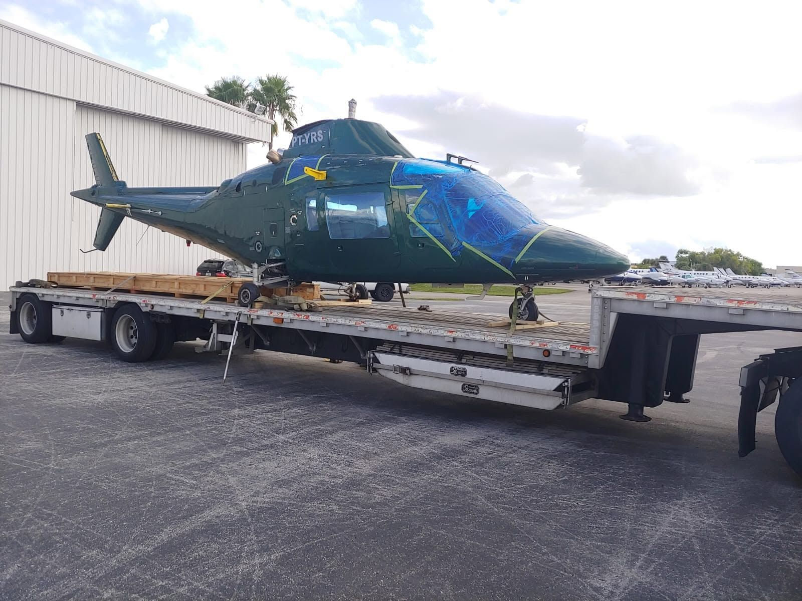 Green helicopter with rotor blades removed, secured on a flatbed trailer and prepared for ground transport outside the hangar.