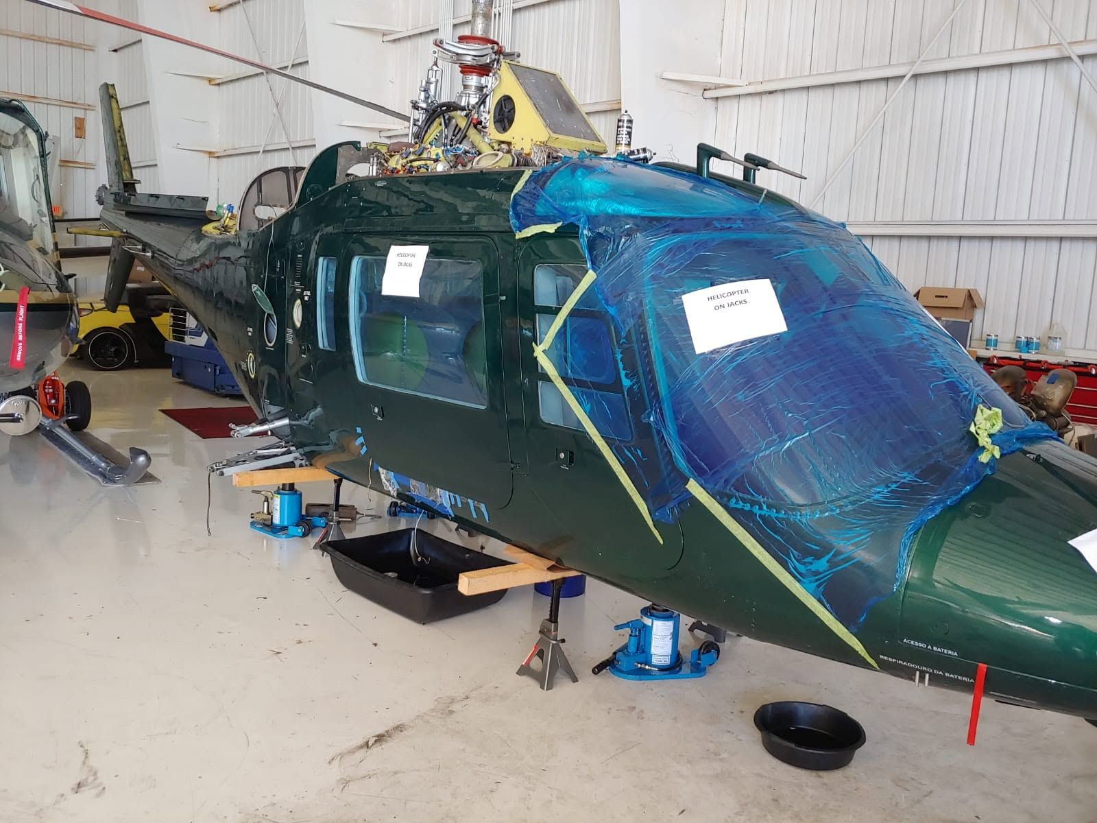 Green helicopter raised on hydraulic jacks for landing gear and rotor system maintenance, with its windshield protected in the hangar.