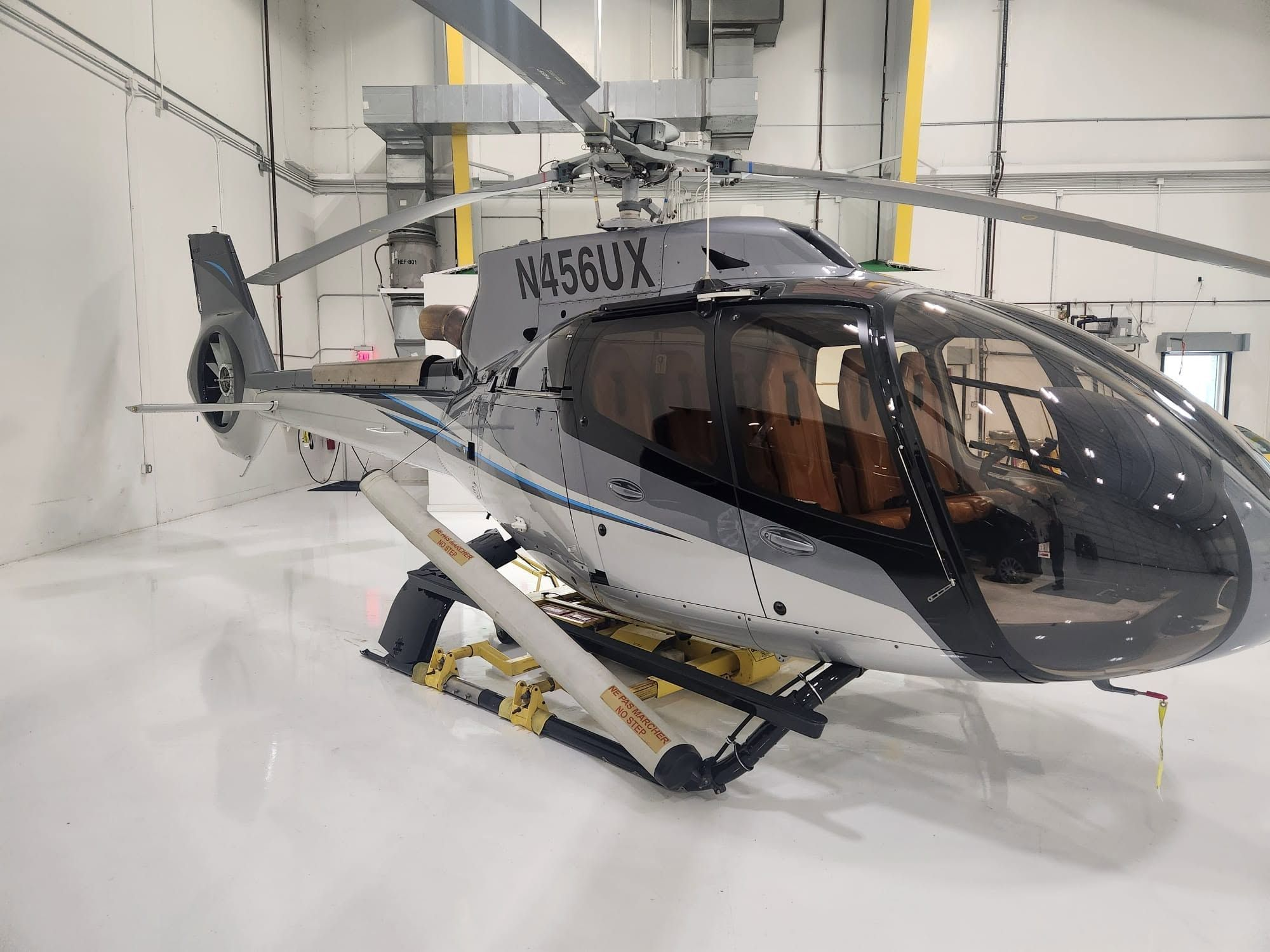Airbus H130 helicopter in gray and silver livery with a tan leather interior, parked in a clean, modern hangar on ground-handling wheels.