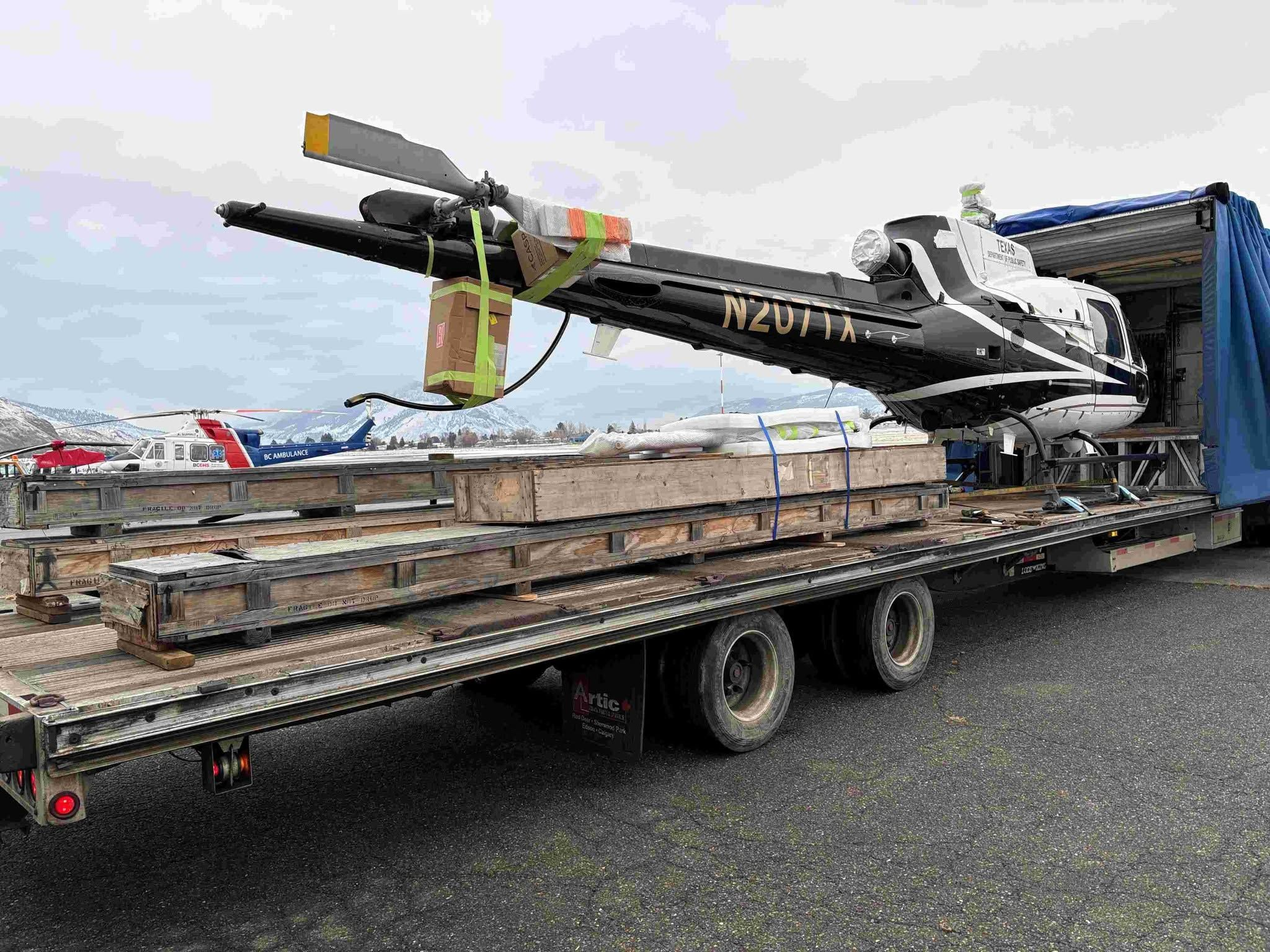 Black and white helicopter loaded on a semi-trailer with its rotor blades packed in wooden crates, ready for long-distance ground transport.