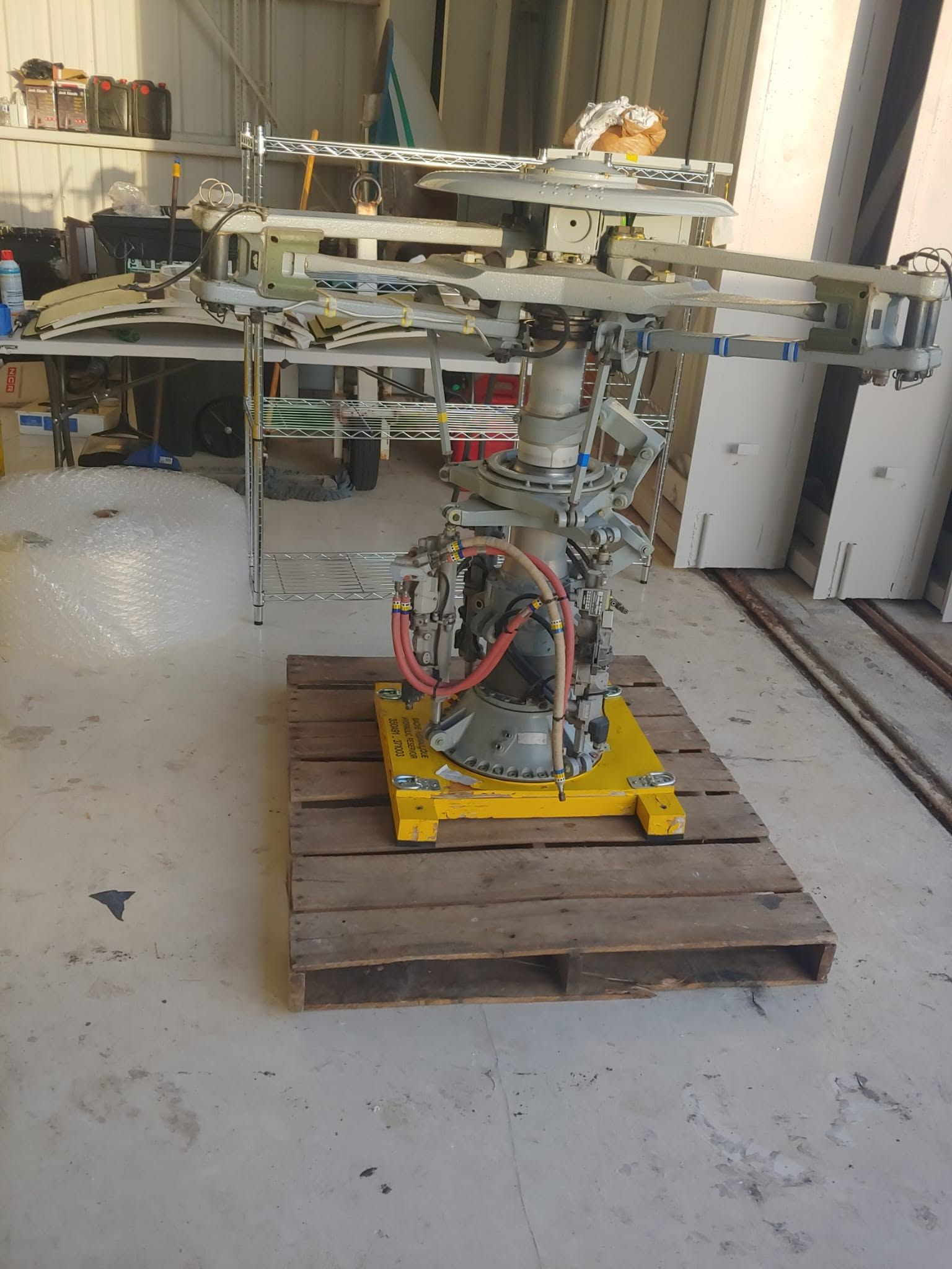 Helicopter main rotor head with mast, swashplate and hydraulic servos, mounted on a yellow transport stand during maintenance.
