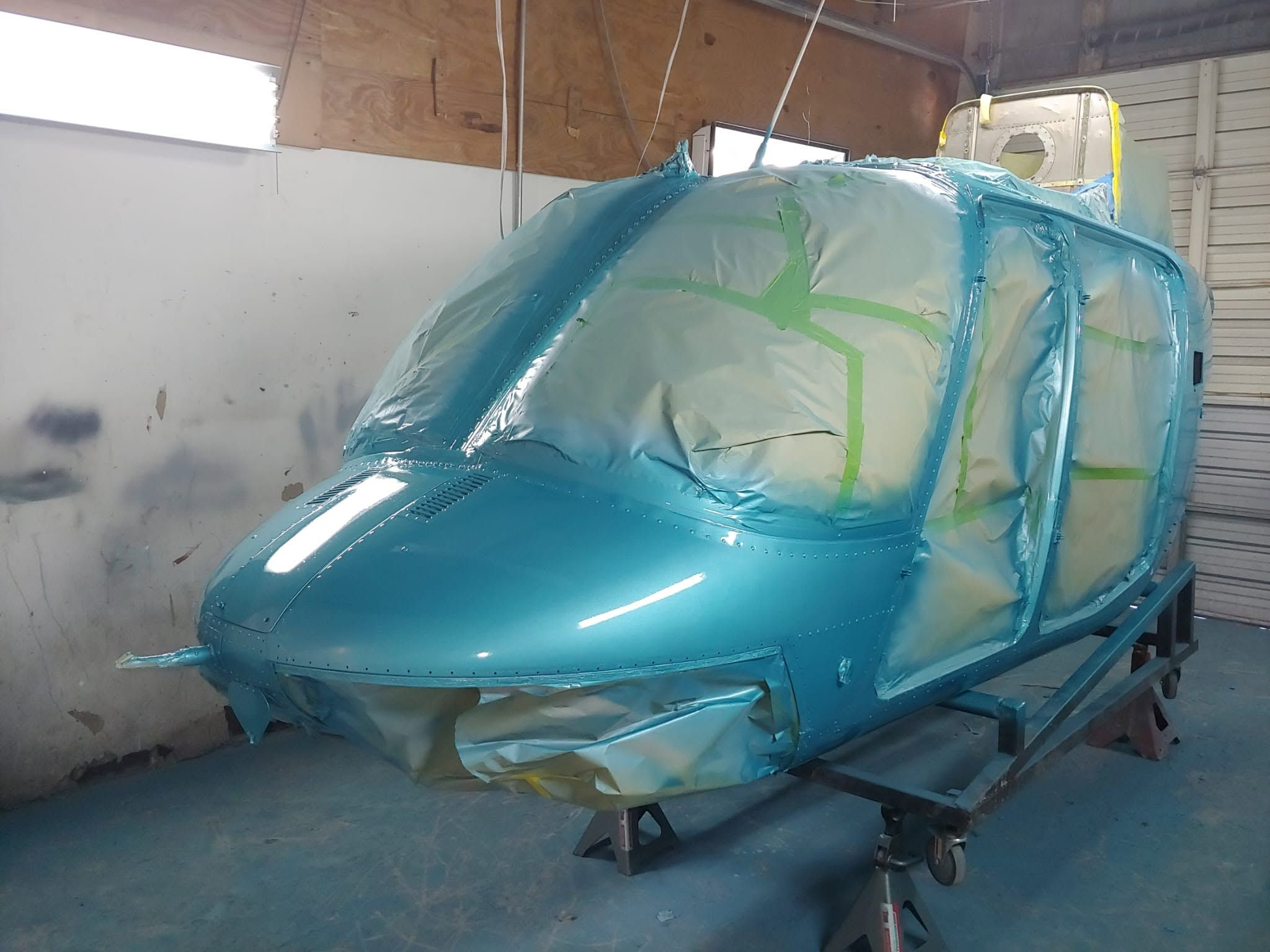 Helicopter fuselage freshly painted in metallic turquoise, with masked windows, mounted on a stand in the paint booth.