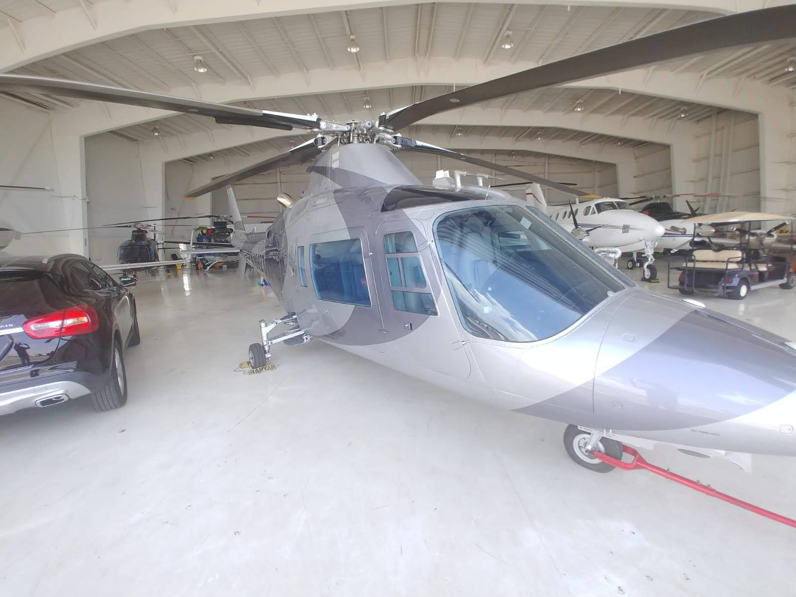 Twin-engine executive helicopter in silver and gray livery, parked inside the hangar with a tow bar attached and other aircraft nearby.