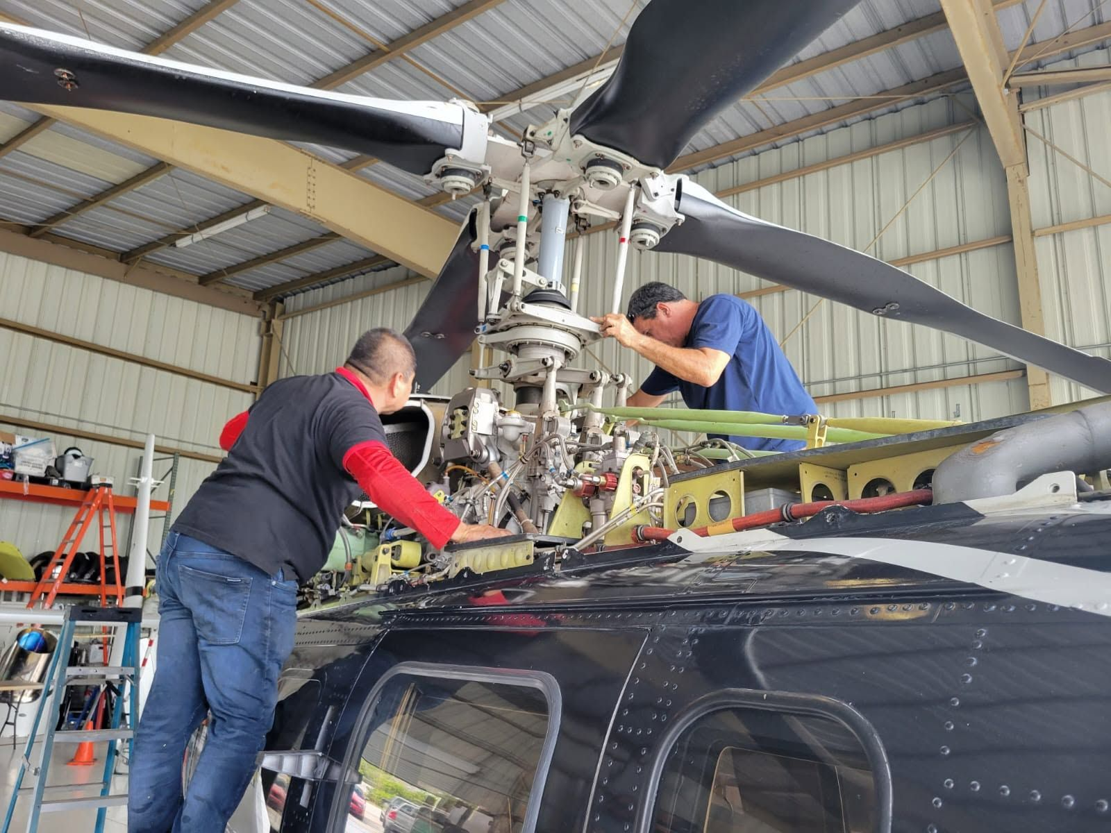 Technicians performing maintenance on a helicopter's main rotor system and flight controls inside the hangar.