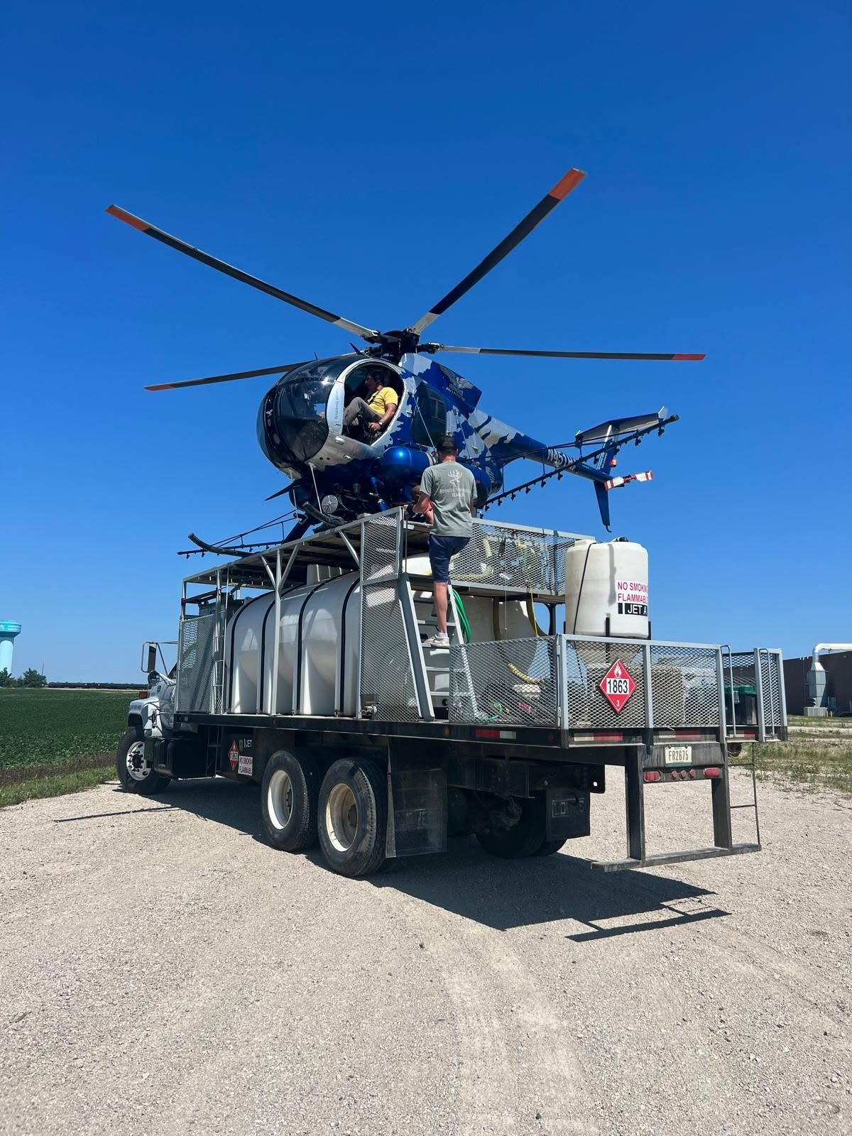 Agricultural spray helicopter with boom sprayers, landed on a support truck carrying Jet A fuel and mixing tanks next to a crop field.